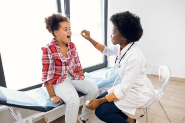 Güzel 10 yaşında melez bir ırk kızı, ağzını açıp boğazını gösteriyor, modern tıp ofisinde genç Afrikalı doktor laringolojisti ziyaret ederken. Boğaz ağrısı, farenjit, bademcik iltihabı..