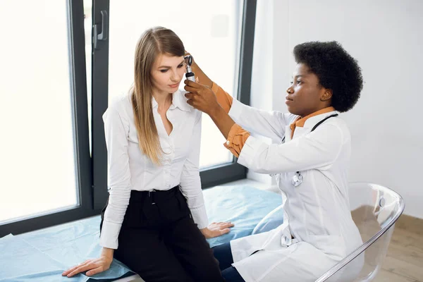 Wanita muda Kaukasia menarik mendapatkan pemeriksaan telinga medis di klinik modern. Praktisi perempuan afro memeriksa telinga pasien, menggunakan otoskop, di kantor dokter. — Stok Foto