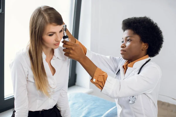 Médico otorrinolaringologista e paciente no hospital moderno. Feminino confiante médico afro-americano fazendo exame de orelha com otoscópio para sua paciente, jovem senhora europeia, sofrendo de dor de ouvido e otite. — Fotografia de Stock