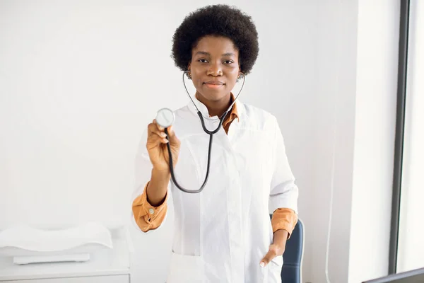 하얀 코트를 입고 미소를 지으며 청진기를 보여 주고 밝은 의학적 배경 위에 실내에 위치한 아프리카 계 미국인 의사 또는 간호사. 의료 및 가정용 의약품. — 스톡 사진
