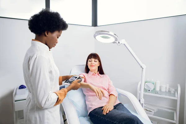 Skin cancer and melanoma prevention. Professional African lady doctor dermatologist examines the patients moles on arm with the help of a modern device for the dematoscopy.