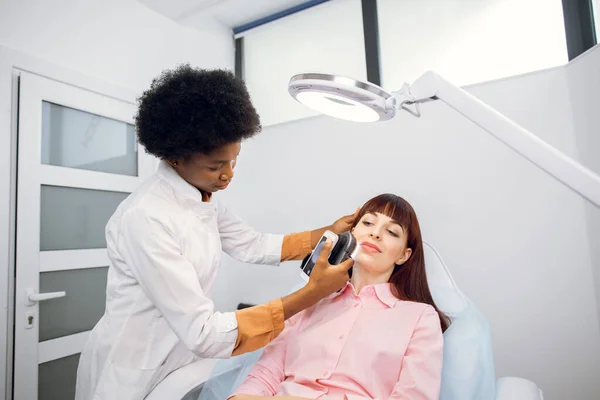Giovane cliente donna sdraiata sul divano in moderna clinica dermatologia, avendo diagnosi della pelle del viso. Professionale giovane dermatologo afro-americano che studia il tipo di pelle utilizzando dermatoscopio. — Foto Stock