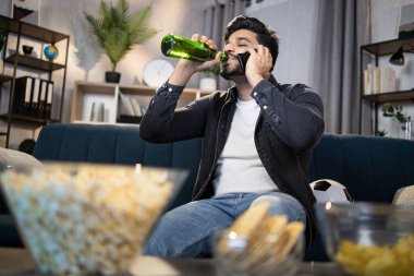 Akıllı telefonu ve birası olan Müslüman adam futbol seyrediyor.