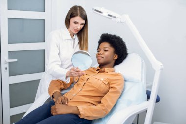 Koltuğunda oturan ve kozmetoloji kliniğinde aynaya bakan güzel bir Afrikalı kadın. Gülümseyen profesyonel güzellik uzmanı mazo terapi ya da cilt bakımından önce cilt prosedürlerini açıklıyor..