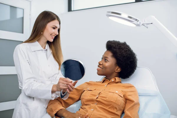 Consulta na clínica de cosmetologia. Médica de beleza feminina conversando com paciente, jovem afro-americana, olhando para o espelho e satisfeita após os procedimentos de beleza. — Fotografia de Stock
