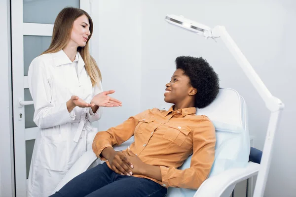 Consulta na clínica de cosmetologia. Médico de beleza profissional feminino conversando com paciente mulher africana muito sorridente e contando boas notícias sobre cuidados e tratamento da pele. Cuidados com a pele e cosmetologia — Fotografia de Stock