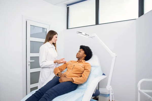 Jovem afro-americana na clínica de cosmetologia de beleza sentada na cadeira médica, enquanto médica vestida de uniforme branco explicando os procedimentos de tratamento da pele. — Fotografia de Stock