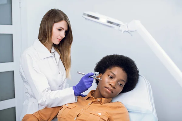 Сучасна концепція технології лікування. Обладнання косметики. Жіночий європейський лікар - косметолог забезпечує процедуру апаратури, кисневу терапію або реактивну очистку для чорношкірого клієнта.. — стокове фото