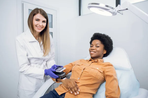 Oncologia, rilevazione del cancro della pelle. Piacevole giovane donna, medico dermatologo guardando la fotocamera con sorriso, mentre il controllo femminile mole pazienti neri a portata di mano con dermatoscopio lente digitale moderna. — Foto Stock