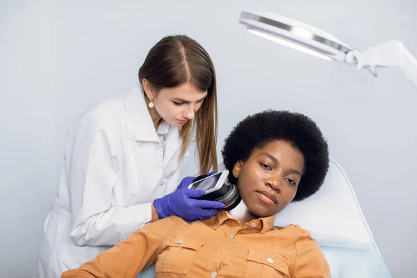 Молода афроамериканська жінка, яка лежить на дивані в сучасній дерматологічній клініці, має діагностику шкіри. Професійна молода жінка - дерматолог, яка вивчає тип шкіри за допомогою дерматоскопа.. — стокове фото