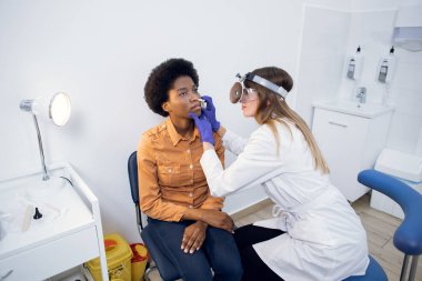 Treatment of nasal diseases. Sinusitis and nasal congestion concept. Caucasian female ENT doctor examines nose of attractive young African girl patient with a medical tool. clipart