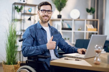 Tekerlekli sandalyedeki adam evde çalışırken başparmağını gösteriyor.