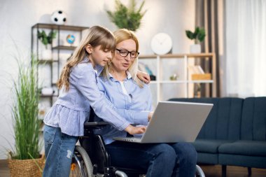 Kızı tekerlekli sandalyedeki anneye dizüstü bilgisayarla sarılıyor.