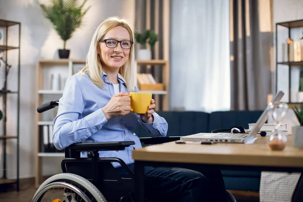 Woman in wheelchair holding cup of coffee and using laptop