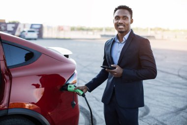 Afrikalı adam elektrikli araba yüklüyor ve akıllı telefon üzerinde çalışıyor.