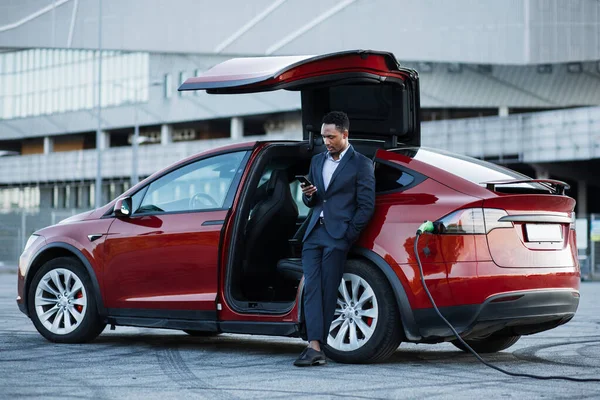 Uomo d'affari che lavora sul cellulare durante la ricarica di auto elettriche — Foto Stock