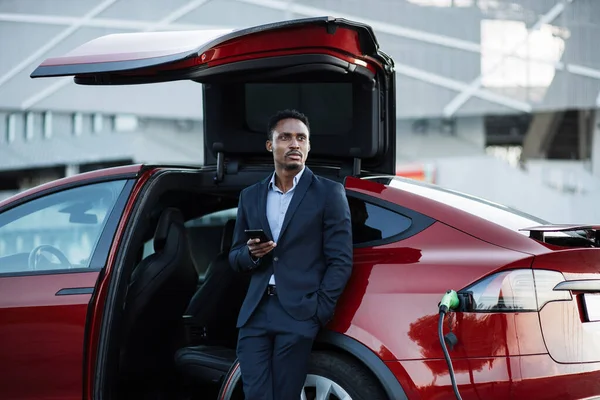 Uomo africano utilizzando il telefono cellulare durante la ricarica auto elettrica — Foto Stock