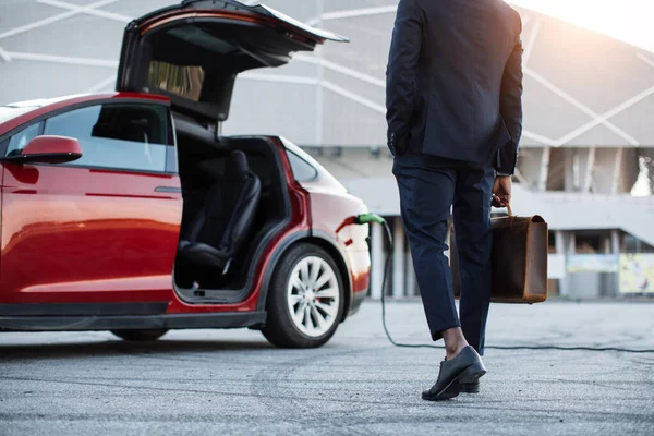 Mężczyzna z walizką stojący w pobliżu elektrycznego samochodu, który ładuje — Zdjęcie stockowe