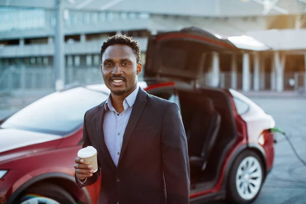 Uomo d'affari che beve caffè mentre l'auto elettrica si ricarica — Foto Stock