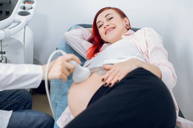 Gülümseyen, neşeli, kızıl saçlı, güzel göbekli beyaz kadın, modern jinekoloji ve doğum kliniğinde ultrason taraması yaptırıyor. Kadın doktorun elleri ultrason taraması yapıyor.