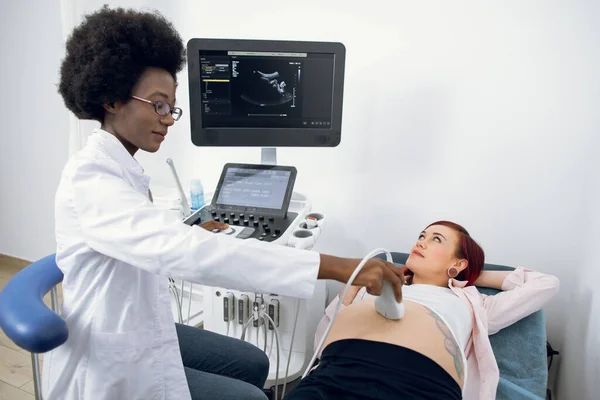 Счастливая молодая беременная белая женщина, проходящая ультразвуковую диагностику в современной клинике, смотрящая на свою черную женщину-врача-акушера, надевая ультразвуковой преобразователь на большой симпатичный живот. — стоковое фото