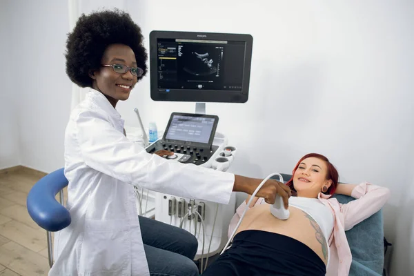 Conceito de ultrassom, obstetrícia. Imagem horizontal de jovem médica afro-americana agradável, olhando para a câmera, durante a realização de procedimento de diagnóstico de ultra-som para a mulher grávida na clínica moderna. — Fotografia de Stock