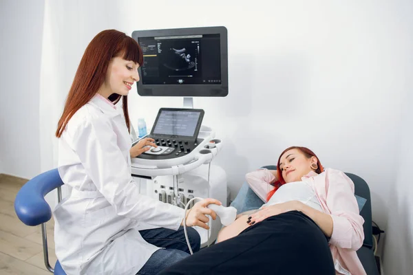 Female happy smiling doctor performs ultrasound examination of her female patient for regular screening of pregnancy at the modern medical office.
