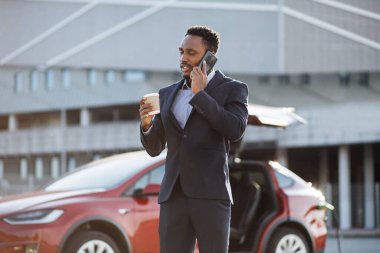 Elektrikli araba şarj olurken iş adamı cep telefonuyla konuşuyor