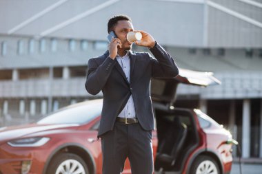 Adam araba şarj olurken kahve içip telefonla konuşuyor.