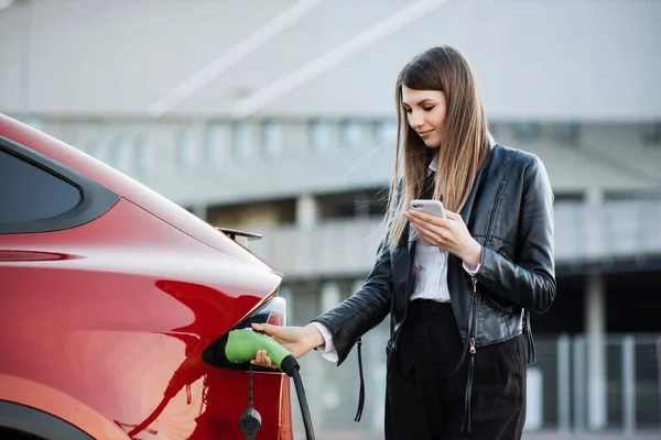 Wanita pengisian mobil listrik dan menggunakan smartphone — Stok Foto