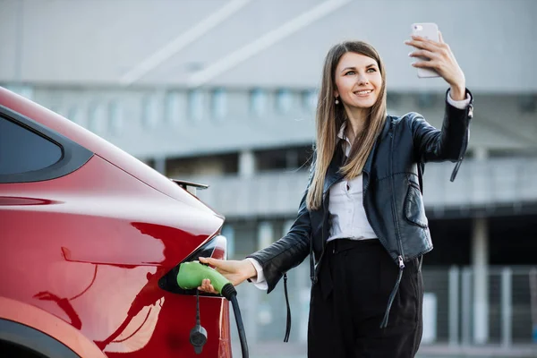 Mujer que tiene videollamada en smartphone mientras carga el coche —  Fotos de Stock