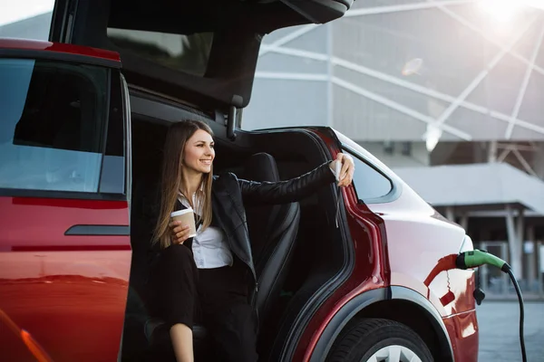 Woman sitting in charging car with mobile and coffee