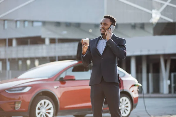 Uomo d'affari che parla sul cellulare mentre l'auto elettrica si ricarica — Foto Stock