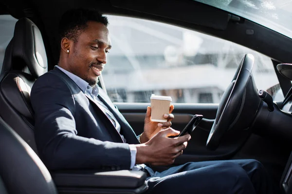 African man in suit drinking coffee and using mobile in car