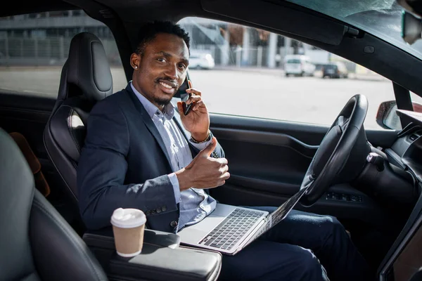 Uomo africano seduto in auto con computer portatile e parlando sul cellulare — Foto Stock