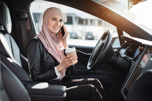 Woman in hijab talking on mobile and drinking coffee in car