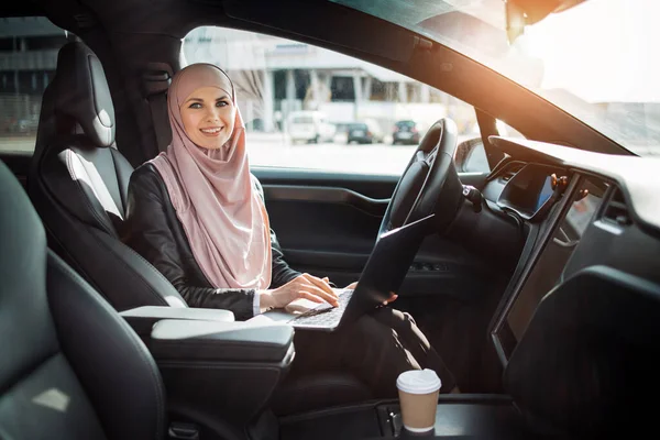 Business lady in hijab sitting in car with laptop on knees