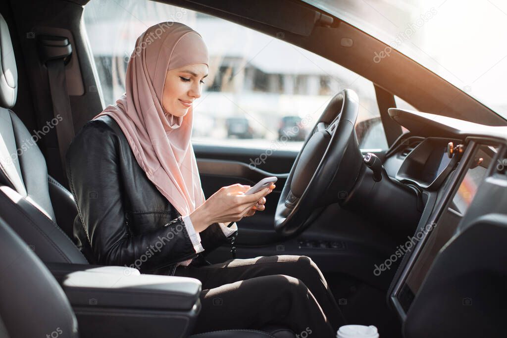 Arabian woman in hijab sitting in car and using smartphone