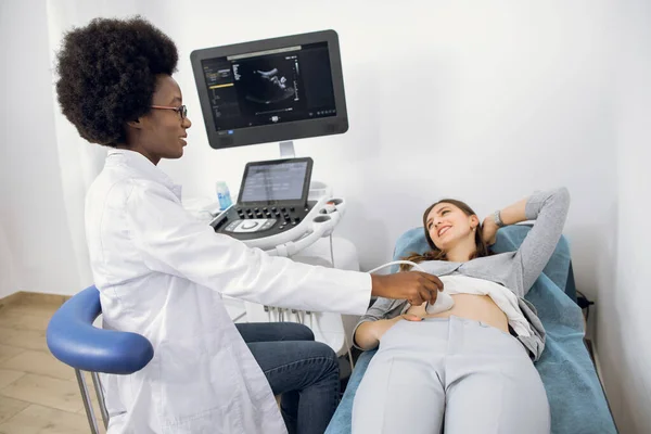 Young African female doctor using ultrasound machine in clinic, makes abdominal ultrasound for young European woman patient. Ultrasound Scanner. Sonography. — Stock Photo, Image