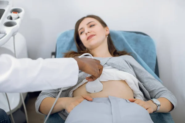 Dispositivo di scansione a ultrasuoni nella mano di un medico afroamericano professionista che esamina giovane paziente caucasica femminile, facendo ecografia ecografia addominale. — Foto Stock