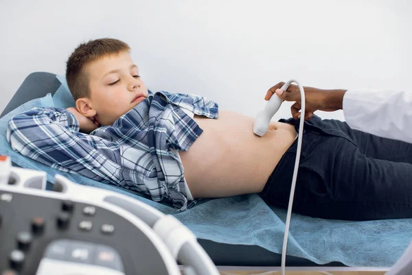 УЗИ ребенка в современной клинике. Женщина-врач сканирует брюшную полость школьника 10-летнего возраста в медицинской клинике. Мальчик лежит на диване и смотрит на сканер USB. — стоковое фото