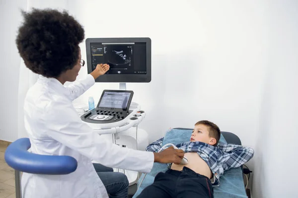 Pediatria e exame médico regular. Médica afro-americana apontando na tela durante a ultra-sonografia dos órgãos abdominais de um menino paciente usando equipamentos modernos na clínica. — Fotografia de Stock