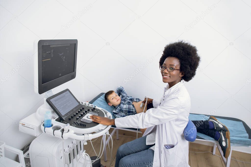Ultrasound examination of the child in modern clinic. Young likable afro american female confident doctor makes an abdominal scan of a cute school boy in a medical clinic.