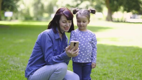 Mère et mignonne petite fille de 3 ans, vêtue de vêtements bleus décontractés, prenant des photos selfie avec caméra téléphonique, faisant un appel vidéo à un ami et saluant, en plein air dans le parc d'été le jour ensoleillé. — Video