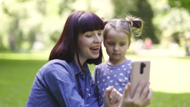 Hezká 30-letá šťastná matka se svou dcerou fotí selfie v parku, usmívá se a baví, ukazuje vítězství mírové znamení na smartphone kamery. Lidé, technologický koncept. — Stock video