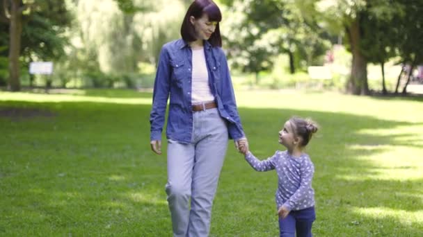 Вид спереди очаровательной симпатичной молодой матери и 4-летней дочери, играющей, держащейся за руки и смотрящей друг на друга, гуляющей в парке красивым солнечным летним утром. — стоковое видео