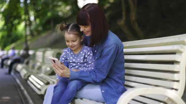 행복 한 어린 어머니와 어린 딸 이 공 원 벤치에 앉아 함께 즐거운 시간을 보내고, 스마트폰에서 게임을 하고 스크린을 스크롤하며, 웃고 즐기는 모습. — 비디오