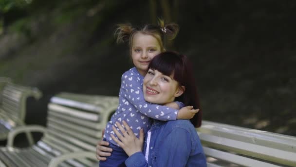아름다운 어린 어머니와 어린 딸은 공원의 야외에서 벤치에 앉아 서로 껴안고 있었다. 행복 한 가족을 붙잡는 것. 행복 한 어머니의 날 기쁨. 느린 동작 4K. — 비디오