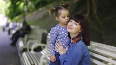Yakın plan, video görüntüleri. Yavaş çekim yumuşak odaklanma. Güzel, genç, beyaz bir anne parkta bankta oturan tatlı küçük kızına sarılıyor. Sevimli kız annesini yanağından öpüyor..