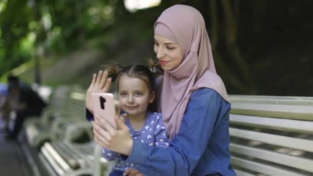 Ganska ung arabisk kvinna i hijab, sitter i parken på bänken tillsammans med sin bedårande lilla dotter, har roligt, vinka till telefonkamera under videosamtal. Människor, familj, teknik. — Stockvideo